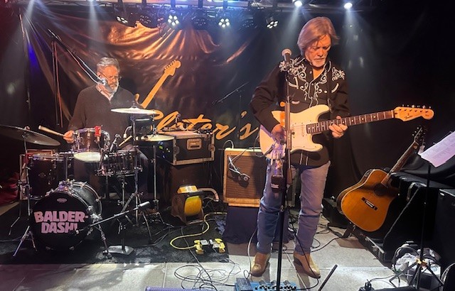 Photo of man playing guitar with drummer in the back.